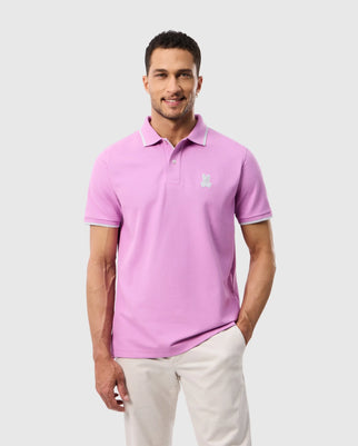 A man stands against a gray background wearing the Psycho Bunny MENS HOUSTON PIQUE POLO SHIRT - B6K604C200 in light purple, featuring a small white logo on his chest, paired with beige pants. The ensemble is accented by the subtle embroidered Bunny design as he smiles with short curly hair.