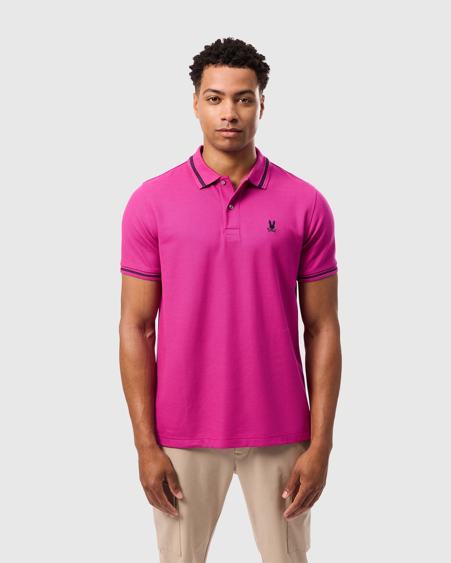 A person wearing a Psycho Bunny MENS RIVIERA PIQUE POLO SHIRT - B6K640C200 in vibrant pink, highlighted with black-trimmed collar and sleeves and adorned with an embroidered Bunny appliqué on the chest. They are standing against a plain light gray background, paired with beige pants for a stylish look.