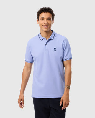 A man stands smiling in the light blue MENS RIVIERA PIQUE POLO SHIRT - B6K640C200 by Psycho Bunny, featuring short sleeves and dark blue trimming on the collar. The Pima cotton shirt showcases a small embroidered Bunny appliqué on the left chest. He has short, curly hair and a tattoo on his left forearm, complementing his dark pants.