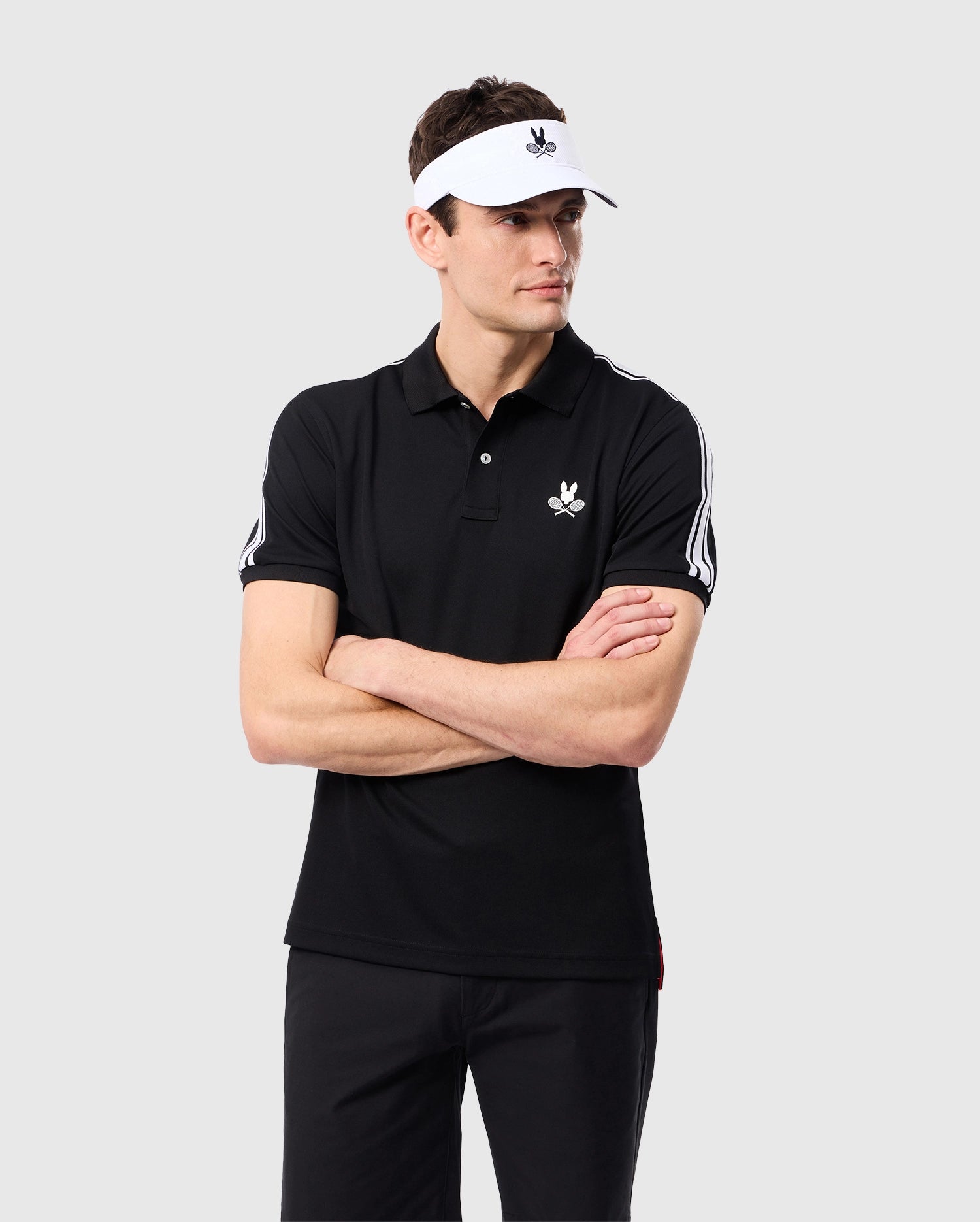 A man stands with his arms crossed, wearing the Psycho Bunny MENS COURTSIDE SPORT POLO - B6K693C200, a black polo featuring white stripes on the sleeves and an embroidered Bunny logo. He pairs it with black polyester pants and completes the look with a white visor. The background is plain and light-colored.