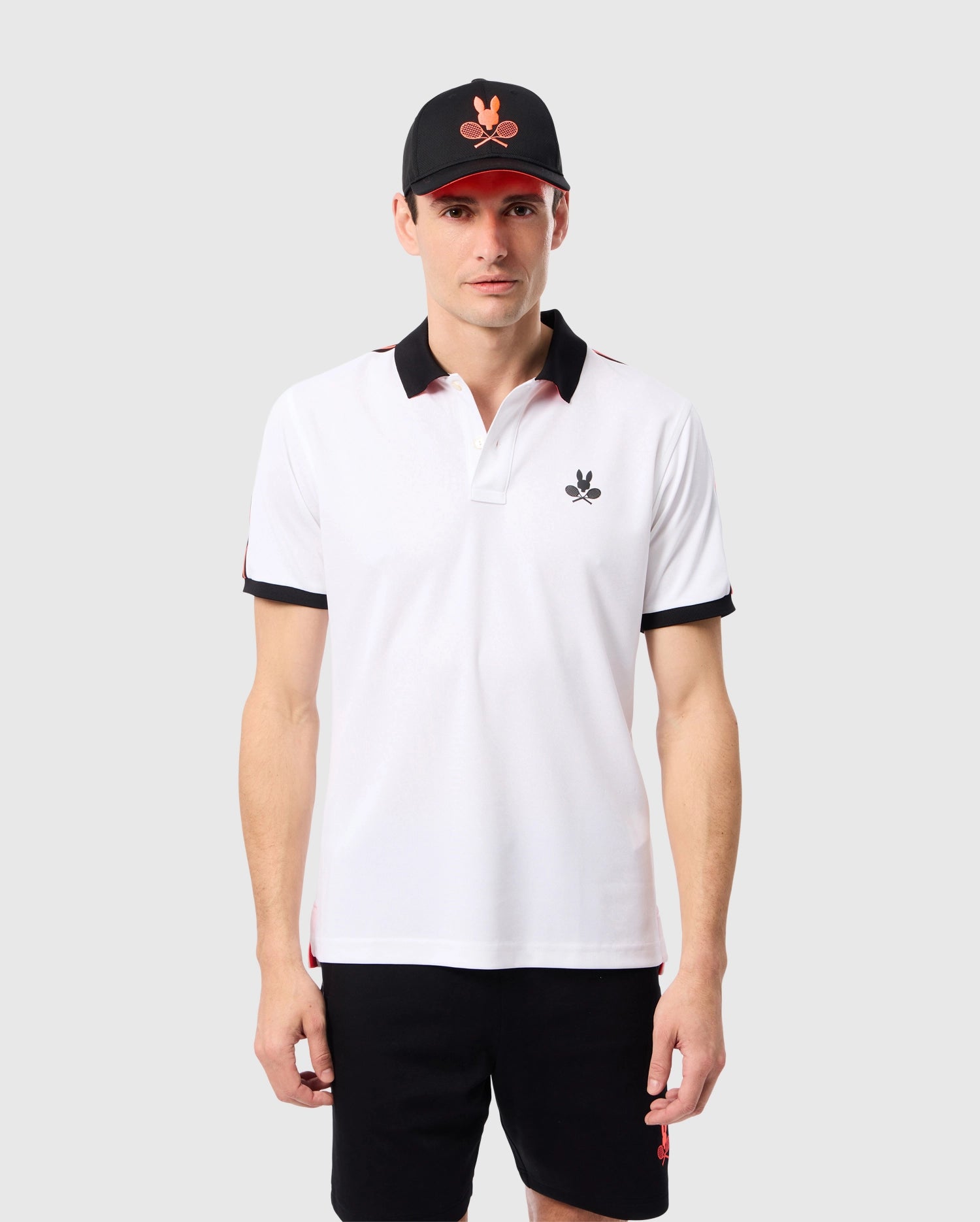 A man stands against a plain background wearing the Psycho Bunny MENS COURTSIDE SPORT POLO - B6K693C200, which has a rib knit collar and black sleeve cuffs. The shirt features an embroidered Bunny logo on the chest. He is also wearing a black cap with a red and white emblem, and black shorts.