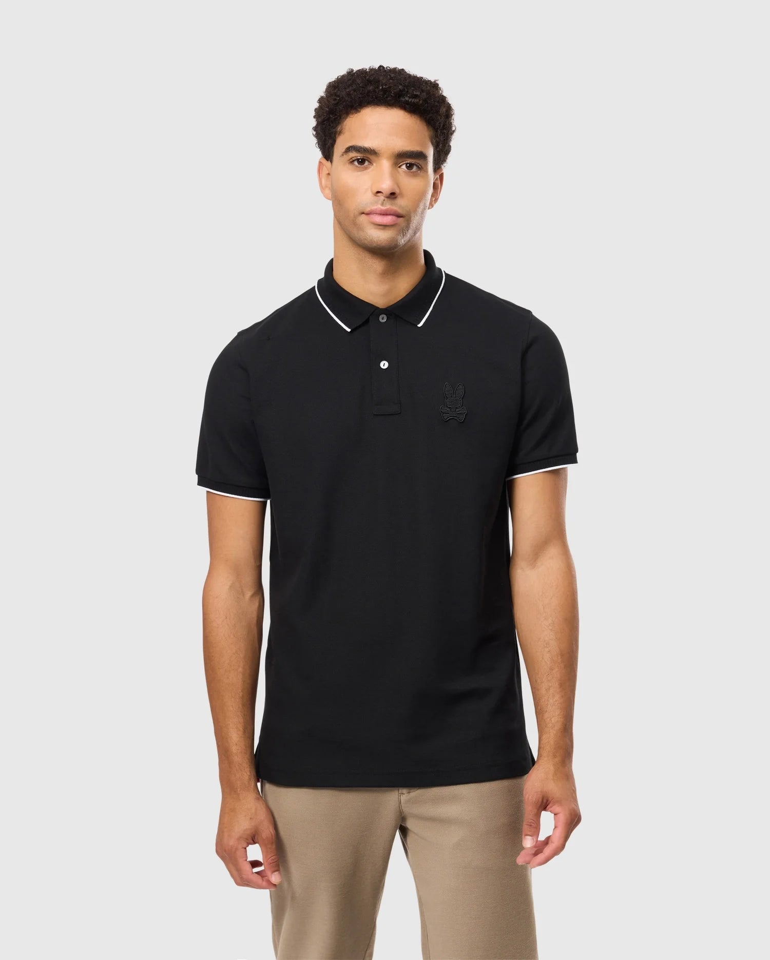 A young man stands against a plain background wearing the Psycho Bunny Mens Kingston Pique Polo Shirt (B6K764D200) in black, featuring white piping on the collar and sleeves. The shirt, crafted from soft Pima cotton, displays an embroidered Bunny patch on the left chest. He has short curly hair and a neutral expression, complemented by beige trousers.