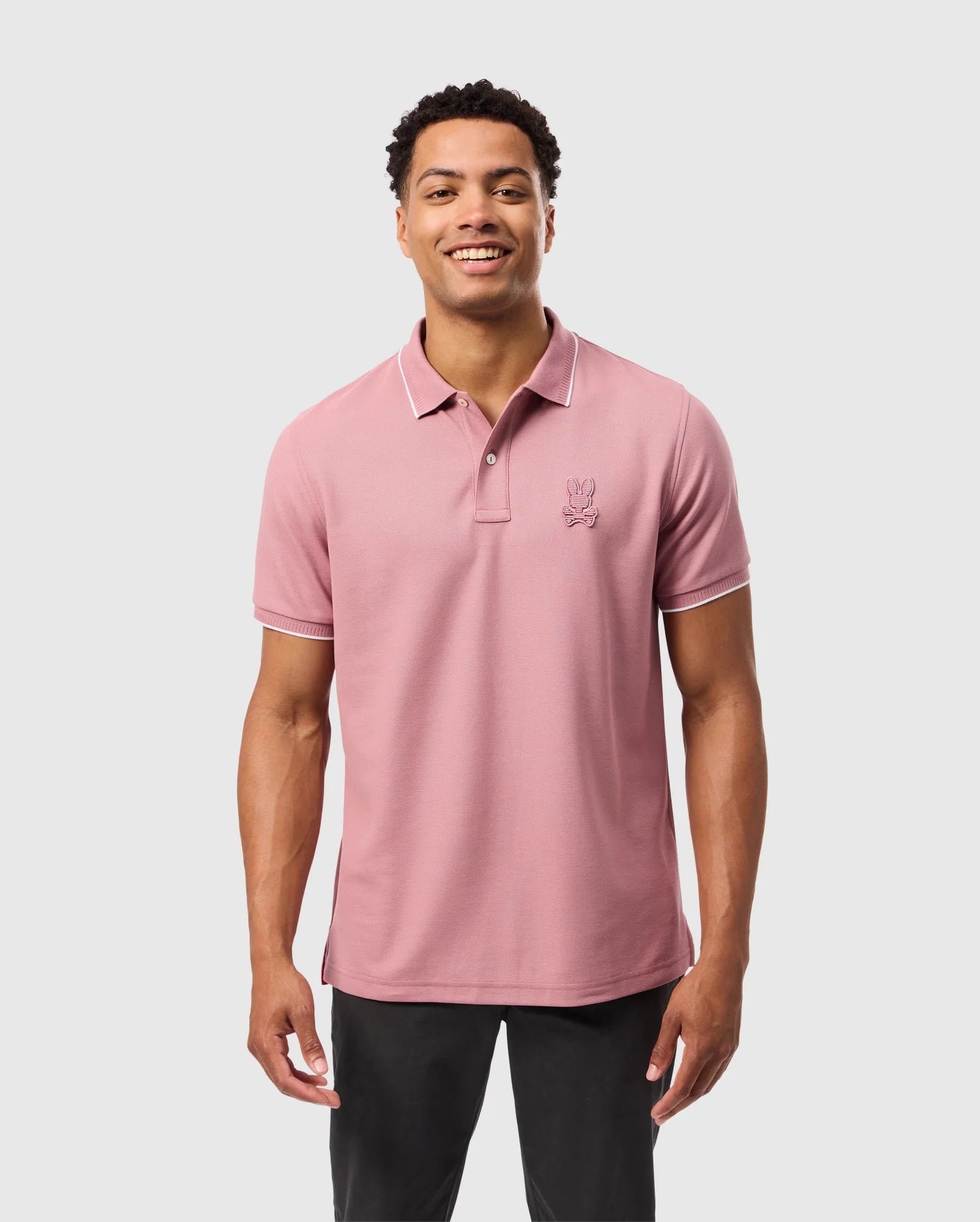 A person smiling against a plain background is wearing a pink Psycho Bunny Men's Kingston Pique Polo Shirt (B6K764D200) made from soft Pima cotton, featuring an embroidered Bunny patch on the chest.