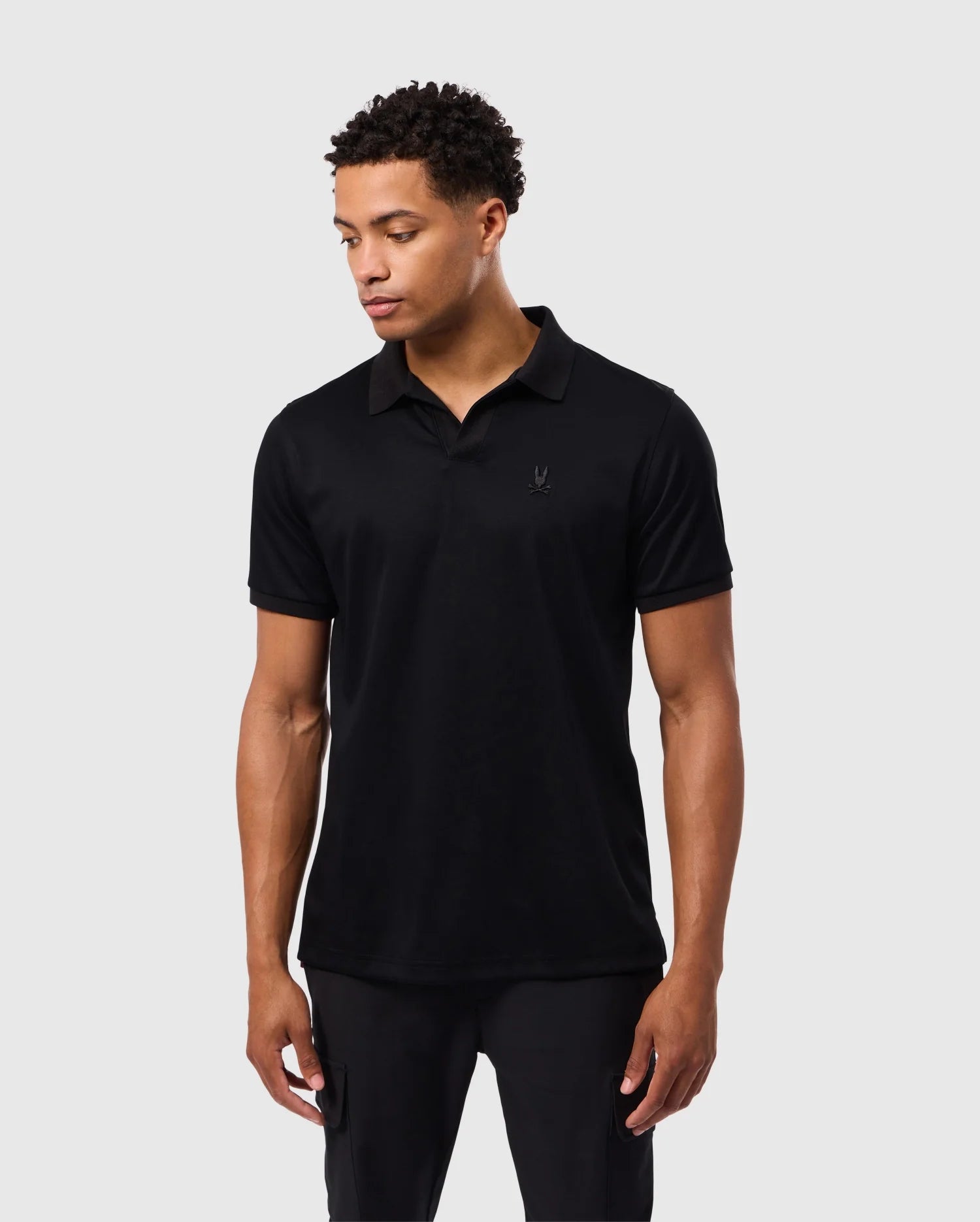 A person with short curly hair is wearing a solid black Psycho Bunny MENS BROADWAY MERCERIZED PIMA POLO SHIRT - B6K891D200 and black pants, standing against a plain light grey background. They are looking slightly downward to their left, with their hands relaxed at their sides.