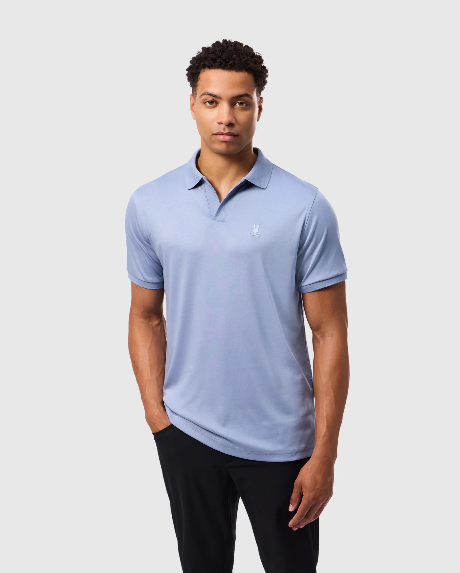 A man with short curly hair is wearing a Psycho Bunny MENS BROADWAY MERCERIZED PIMA POLO SHIRT, light blue in color, and black pants. He has one hand in his pocket and is looking directly at the camera against a plain light gray background.