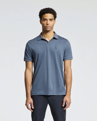 A man stands wearing a Psycho Bunny Men's Broadway Pima Interlock Johnny Collar Polo, showcasing its durable luster and small embroidered logo on the left chest, paired with dark pants against a light, plain background.