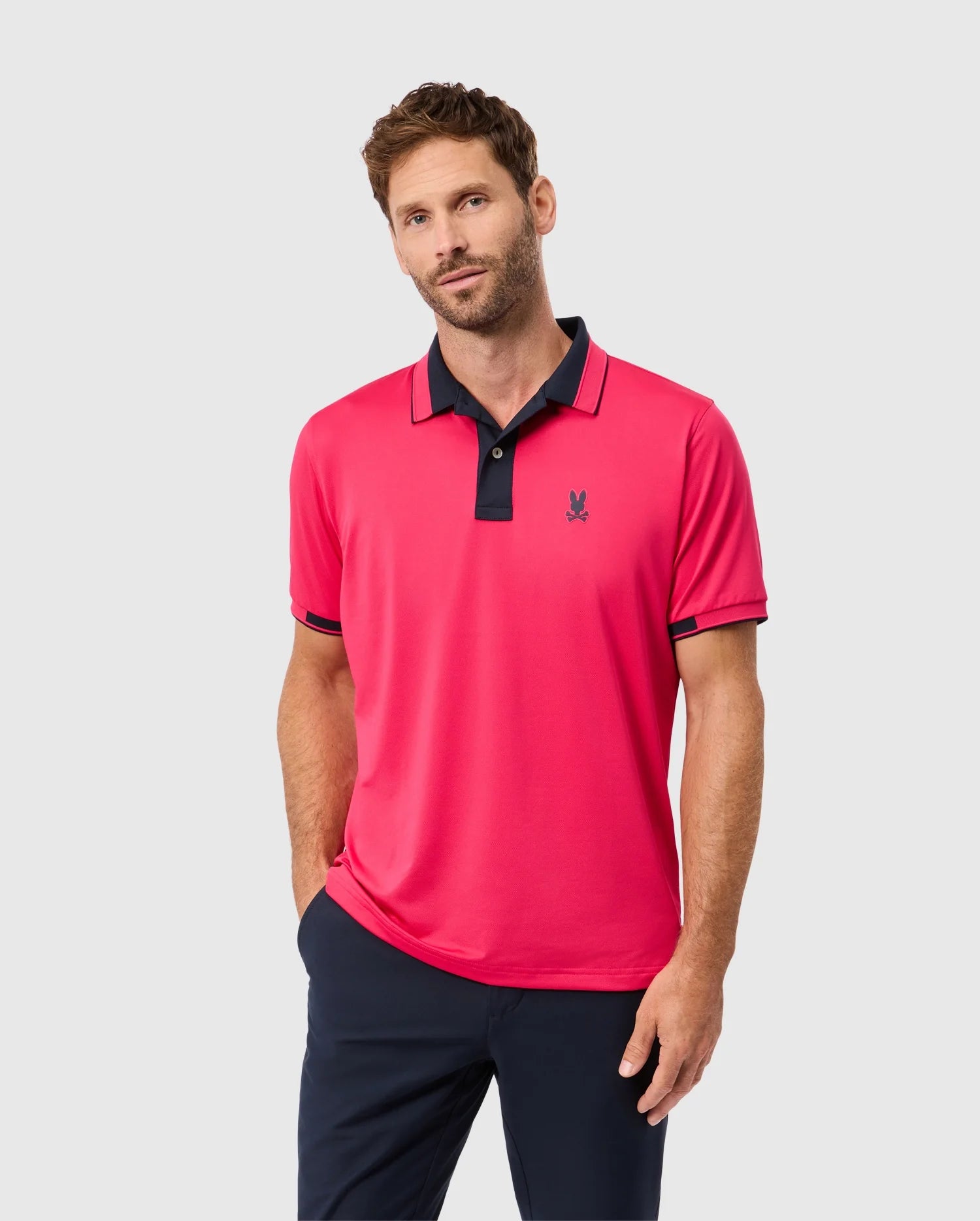 A man with short brown hair and a beard is wearing a Psycho Bunny MENS BELLEVUE SPORT POLO SHIRT - B6K905D200, which is bright pink with black trim on the collar and sleeves, paired with dark pants. He stands against a plain white background, looking at the camera with a neutral expression and has one hand in his pocket.