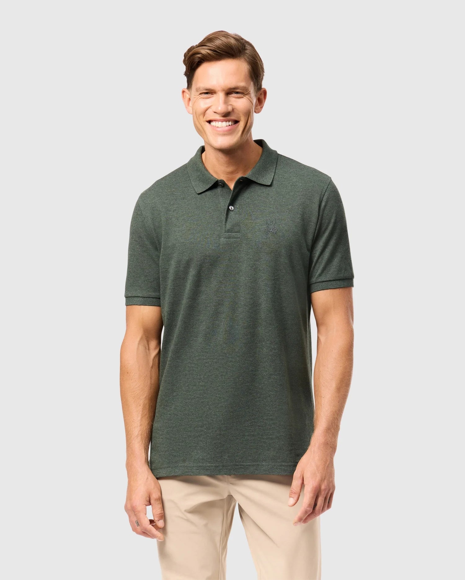 A smiling man wearing a Psycho Bunny MENS JAMES PREMIUM PIQUE POLO SHIRT in dark green and beige pants stands against a plain grey background. He has short, styled brown hair and is looking directly at the camera with his arms casually at his sides.