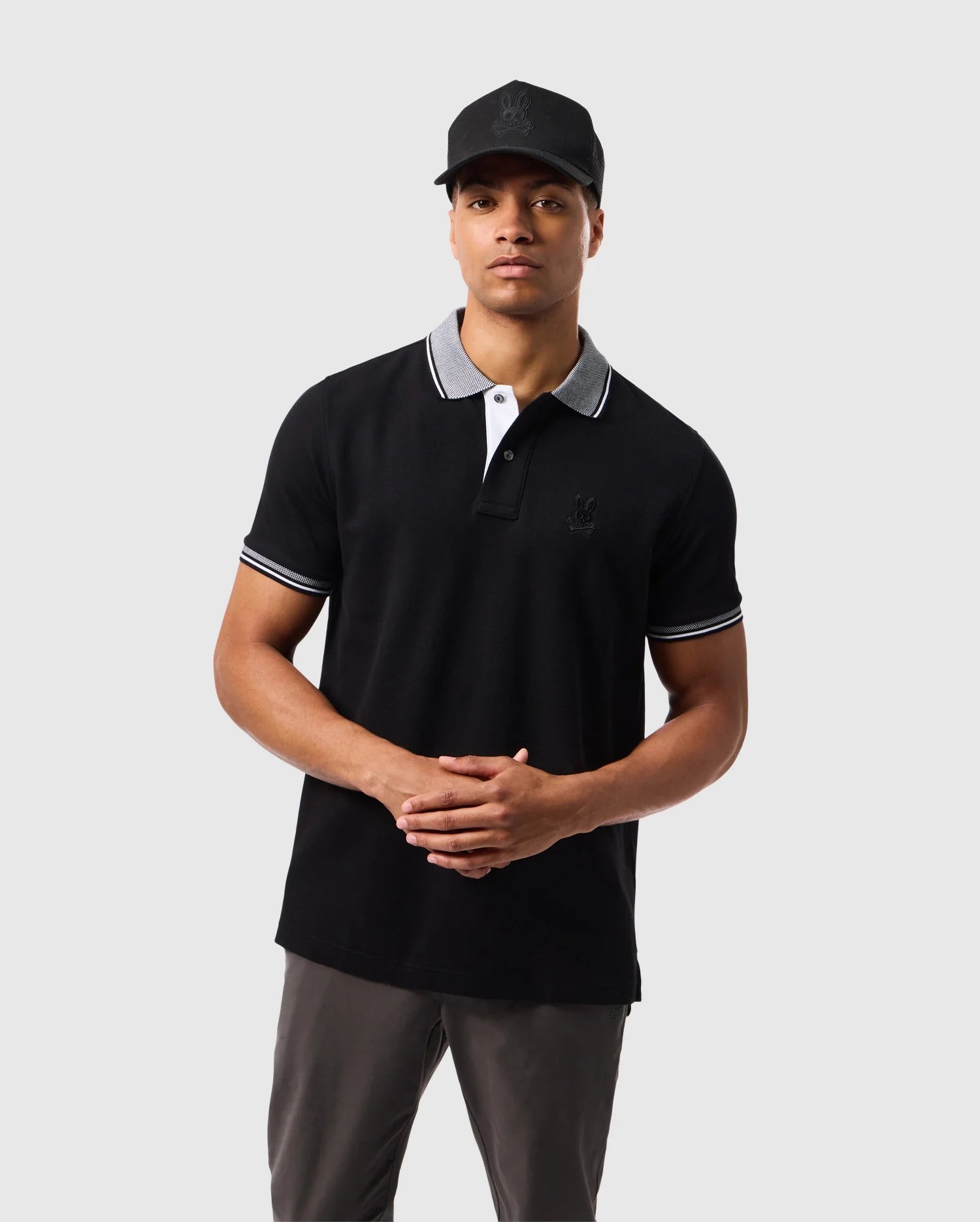 A man stands against a plain background wearing a black MENS MALTON PIQUE POLO SHIRT - B6K950D200 by Psycho Bunny, featuring white collar and sleeve trim. He pairs it with khaki pants and a black cap. The Pima cotton polo enhances his look as he clasps his hands in front of him, looking directly at the camera.