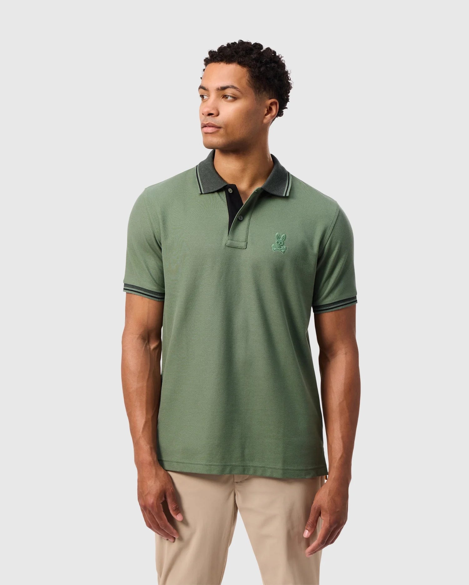 A man is looking to his side while wearing the Psycho Bunny MENS MALTON PIQUE POLO SHIRT - B6K950D200, crafted from soft pima cotton. The shirt features a subtle embroidered logo on the chest and boasts a black collar. It is paired with light khaki pants against a plain white backdrop.