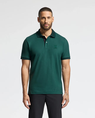 A man with short hair and a beard is wearing a high-quality men's MENS MALTON PIQUE POLO - B6K950D200 by Psycho Bunny in green, paired with black pants. He stands against a plain white background, looking straight ahead.