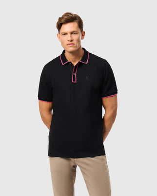 A person wearing the Psycho Bunny MENS KENT PIQUE POLO SHIRT - B6K951D200, a stylish black polo with pink trim, paired with beige pants, stands against a plain gray background. This comfortable polo features a small embroidered emblem on the chest, seamlessly combining elegance and ease.