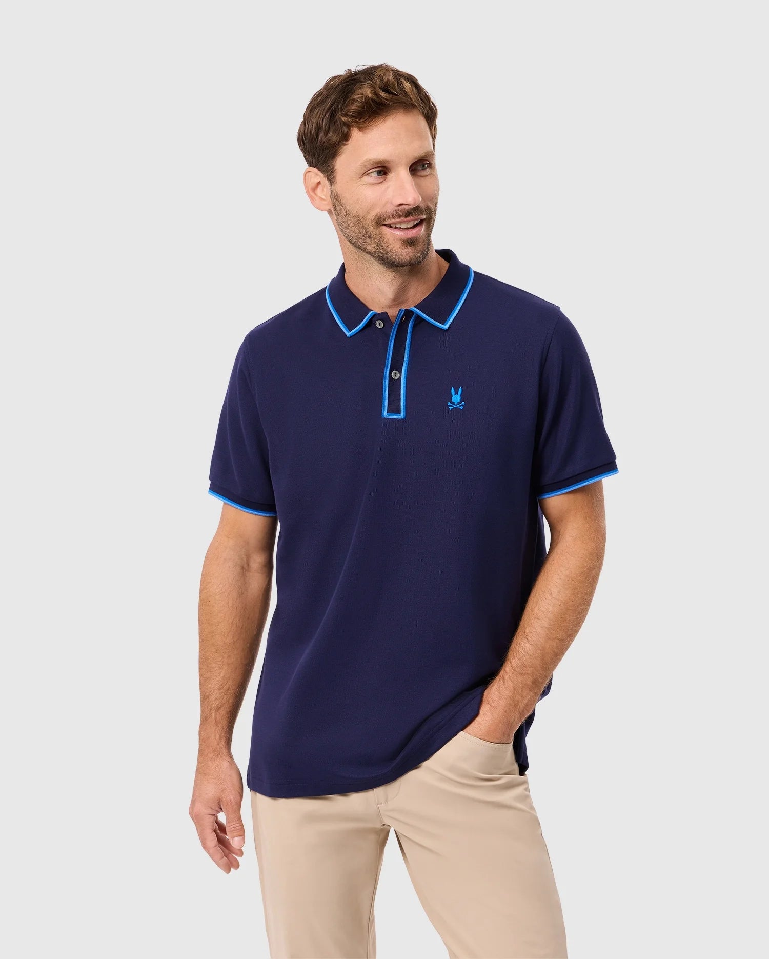 A man with short brown hair and a beard wears the Psycho Bunny MENS KENT PIQUE POLO SHIRT in navy blue, featuring light blue trim and made of comfortable Pima cotton. Matched with beige pants, he smiles against a light gray background with one hand in his pocket.