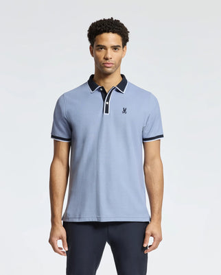 A man in a Psycho Bunny light-blue MENS KENT PIQUE POLO - B6K951D200 with dark trim and a chest emblem stands facing forward against a plain background. The shirt is made of soft Pima cotton. He has short curly hair and wears dark pants.