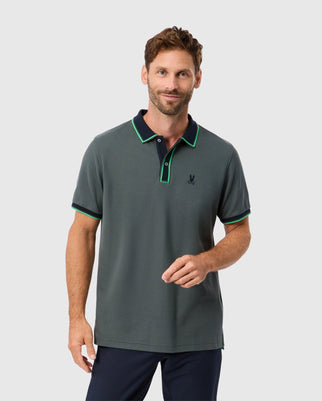 A man with short brown hair and a beard is wearing the stylish MENS KENT PIQUE POLO SHIRT - B6K951D200 by Psycho Bunny, featuring gray fabric with green trim and a small chest emblem. This comfortable polo gives him a relaxed expression as he stands against a light gray background.