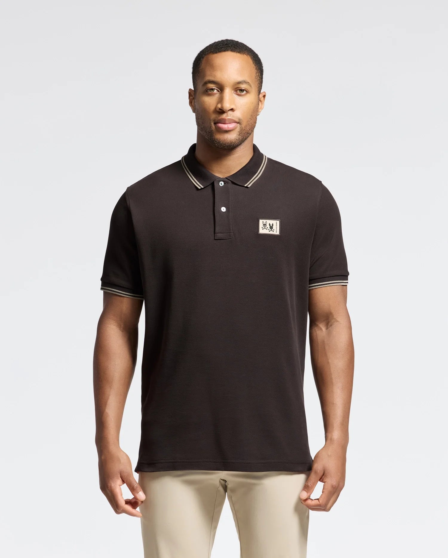 A man stylishly pairs beige pants with a Psycho Bunny Men's Monroe Pique Polo (B6K964D200) featuring Mother-of-Pearl buttons, an embroidered chest logo, and a ribbed collar with lighter outline details against a plain background.