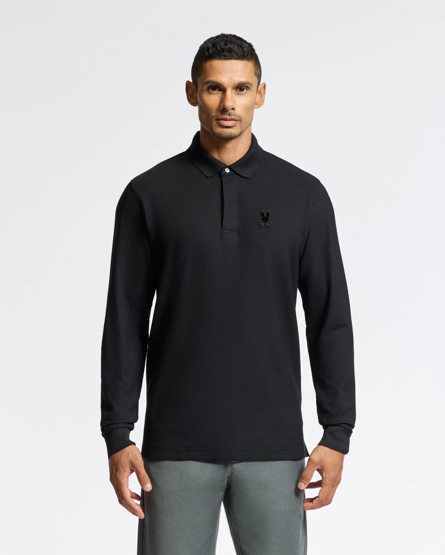 A man models the stylish Psycho Bunny MENS BRANSON LONG SLEEVE PIQUE POLO - B6M177E200, its subtle emblem adding elegance. The lightweight, breathable design pairs perfectly with his gray pants, set against a plain white backdrop.