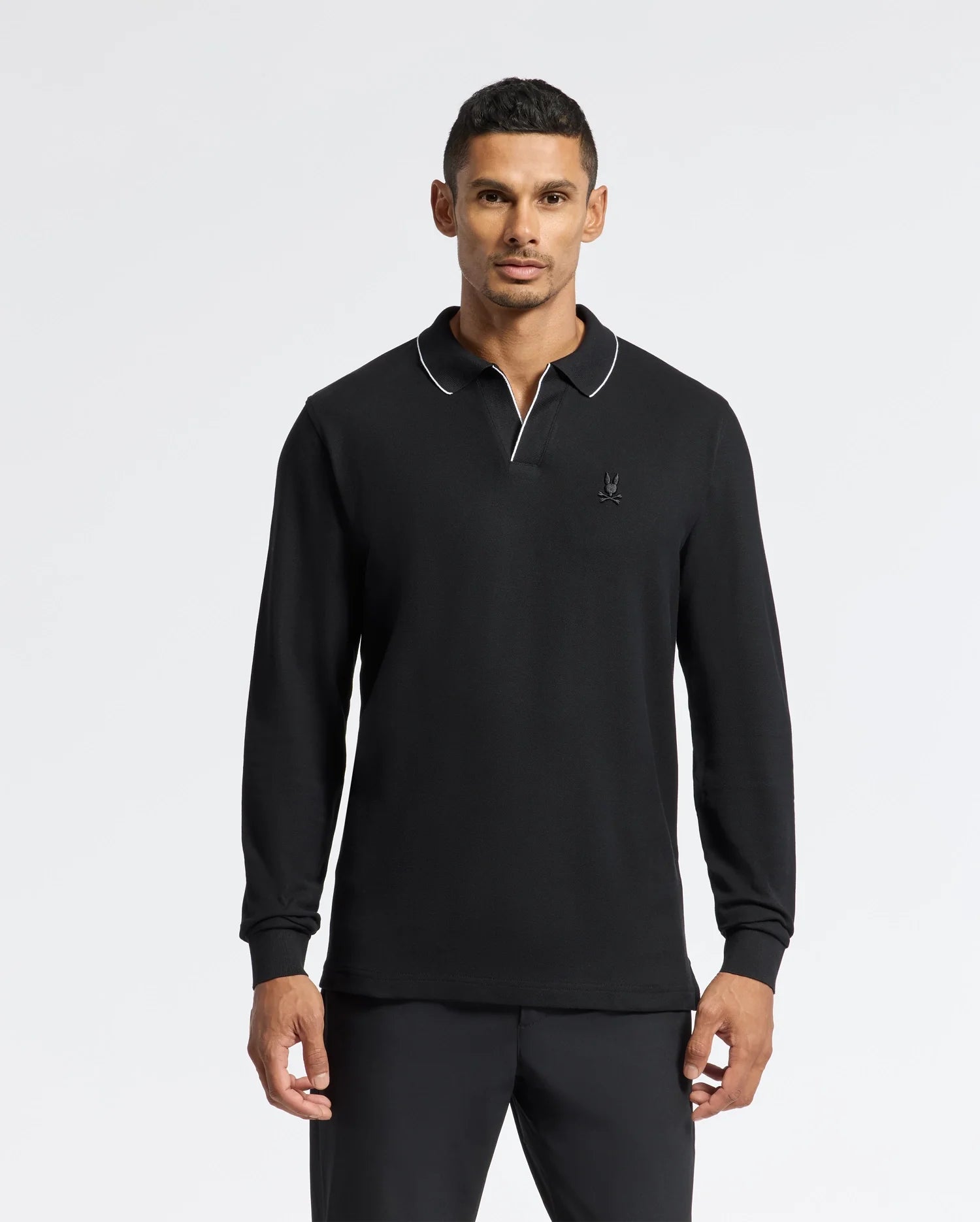 A person with short, dark hair stands against a plain background, wearing the Psycho Bunny Men's Creede Pique Johnny Collar Polo (B6M265E200). This black long sleeve polo is accented with white piping on the collar and made from soft Pima cotton. It features a small emblem on the chest.