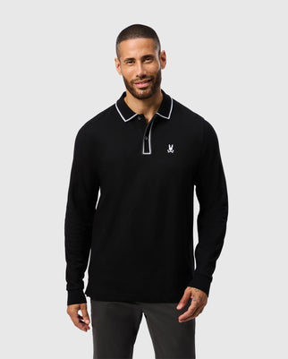 A man with short hair and a beard is wearing a Psycho Bunny Kent Long Sleeve Pique Polo in black, exuding quiet luxury with its white trim and a small embroidered logo on the chest. He stands smiling against a plain light gray background.