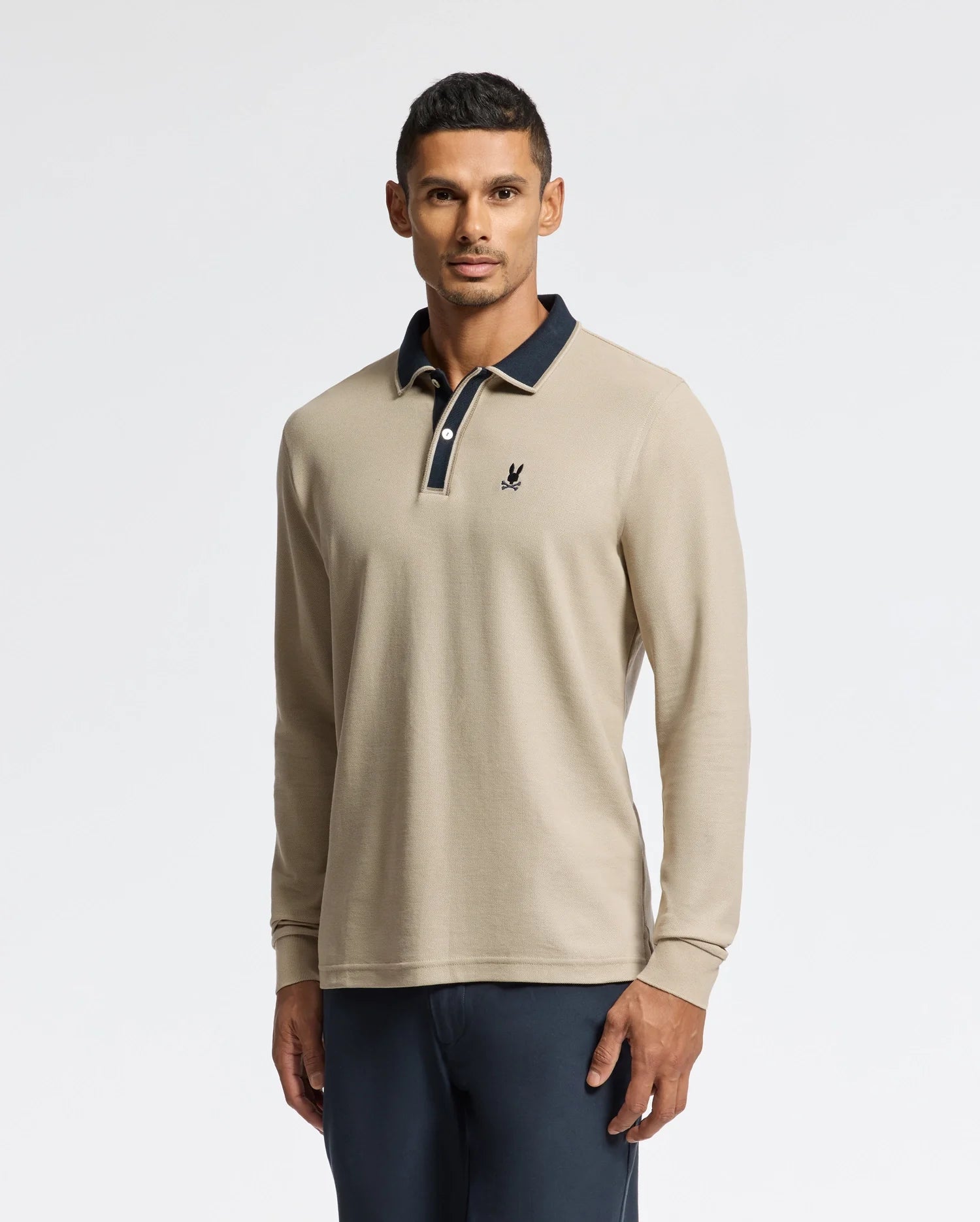 Against a plain background, an individual exemplifies understated elegance in the Psycho Bunny Men's Kent Long Sleeve Pique Polo (B6M935D200) with a black collar and emblem. The sophisticated beige top is seamlessly paired with dark pants for a polished look.