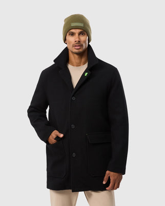 A man wearing the Psycho Bunny MENS STAFFORD WOOL BLEND COAT - B6N551D200, with his left hand in the pocket, stands against a plain background. He has a light brown knit beanie on his head and a beige shirt underneath the stylish men's winter coat, which features a small green pin on the left lapel.