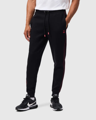 A person adorned in the MENS TRAVIS TRACK PANT - B6P143D200 by Psycho Bunny, featuring a comfy cotton-blend material in black with pink piping for a regular fit, stands against a plain background. Their ensemble is completed with chic black and white sneakers.