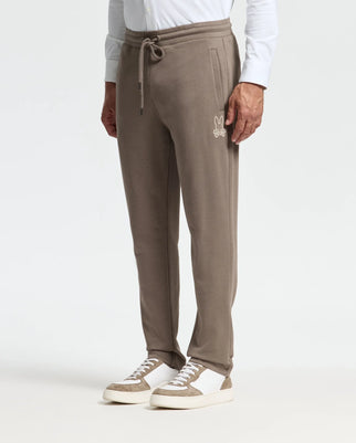 A person wearing the MENS ALEXANDER SWEATPANT - B6P155E200 by Psycho Bunny, featuring a taupe color, relaxed fit, drawstring waist, and rabbit logo. They have paired it with white sneakers and a white long-sleeve shirt. Their hands rest at their sides against a plain white background.