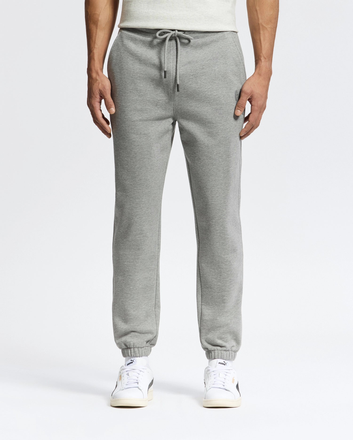 The lower half of a person is shown, wearing Psycho Bunny's MENS ELDORADO BRUSH BACK JOGGER - B6P285E200 in gray with an elasticized waist and drawstring. Hands are in pockets, and white sneakers complete the outfit against a plain background.