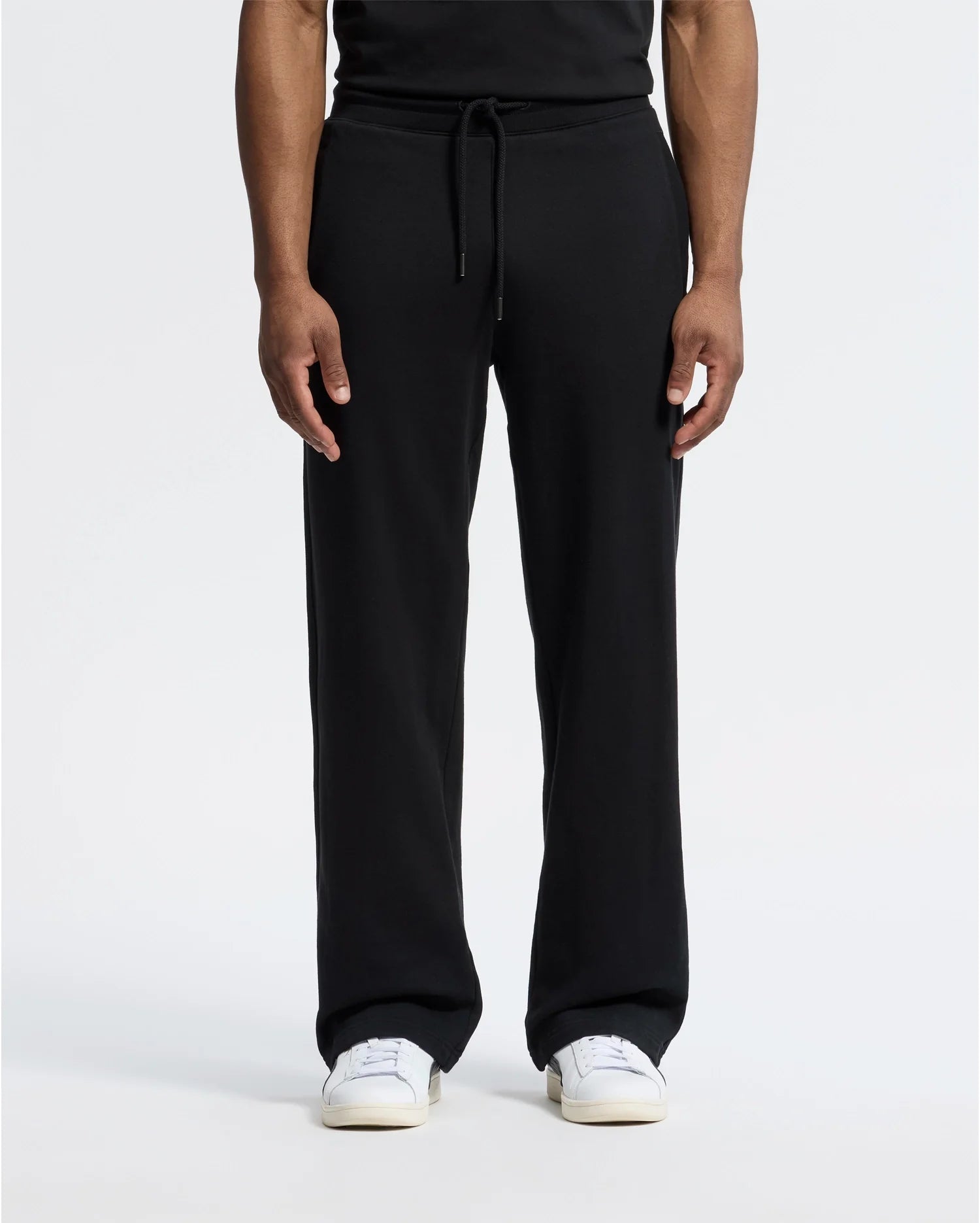 A man is wearing Psycho Bunny's black MENS LYNX WIDE LEG CLASSIC FRENCH TERRY SWEATPANT - B6P379E200 with adjustable drawstring waist, paired with white sneakers, against a plain background. Only his lower body is visible.