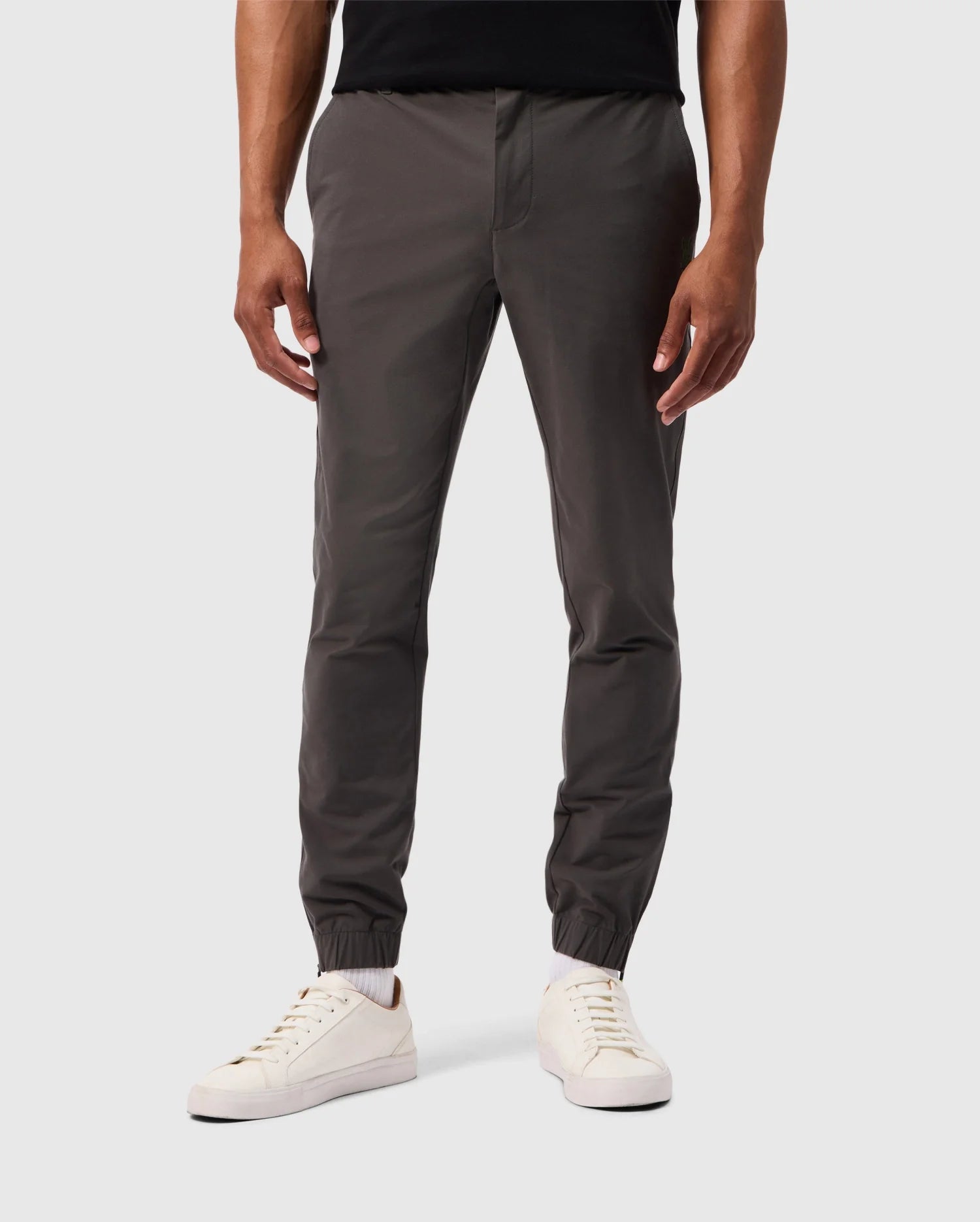 A person wearing dark gray MENS MADISON COMMUTER PANTS - B6P395Z1WB from Psycho Bunny with elastic cuffs stands against a plain white background. Their hands are relaxed by their sides, and they are pairing the pants, featuring a detachable Bunny keychain, with white sneakers and a black top.