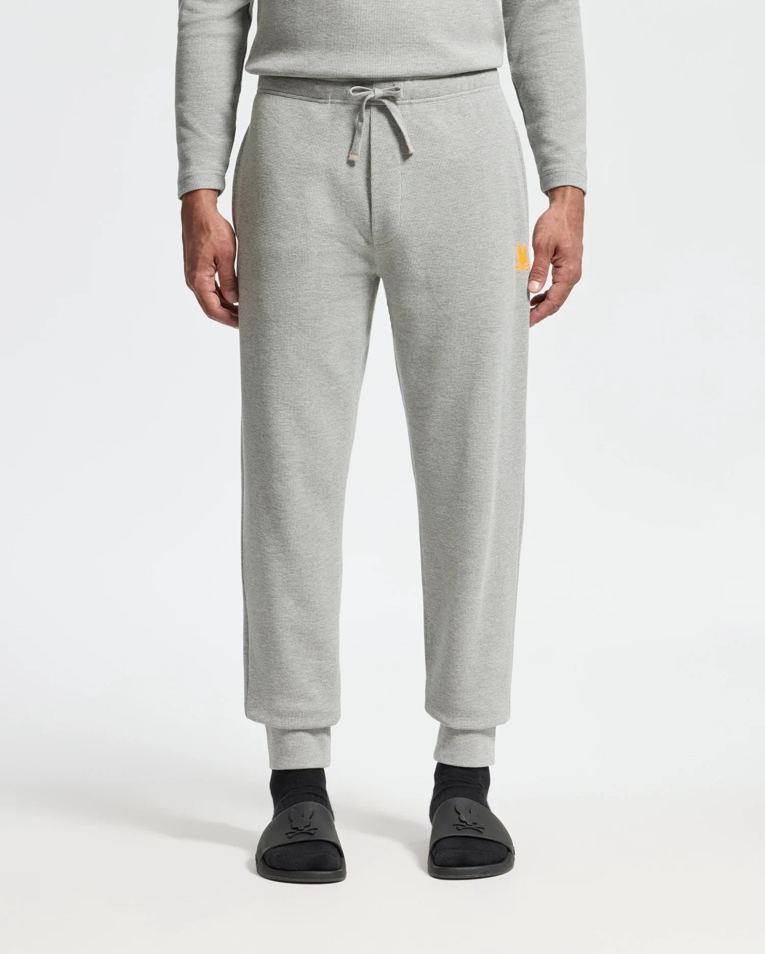 A person is wearing the Psycho Bunny MENS VAIL WAFFLE LOUNGE PANT in light gray, featuring a drawstring waist, paired with black shoes that have simple designs. The matching gray top enhances the sophisticated look against the plain white background, presenting a casual yet refined style.