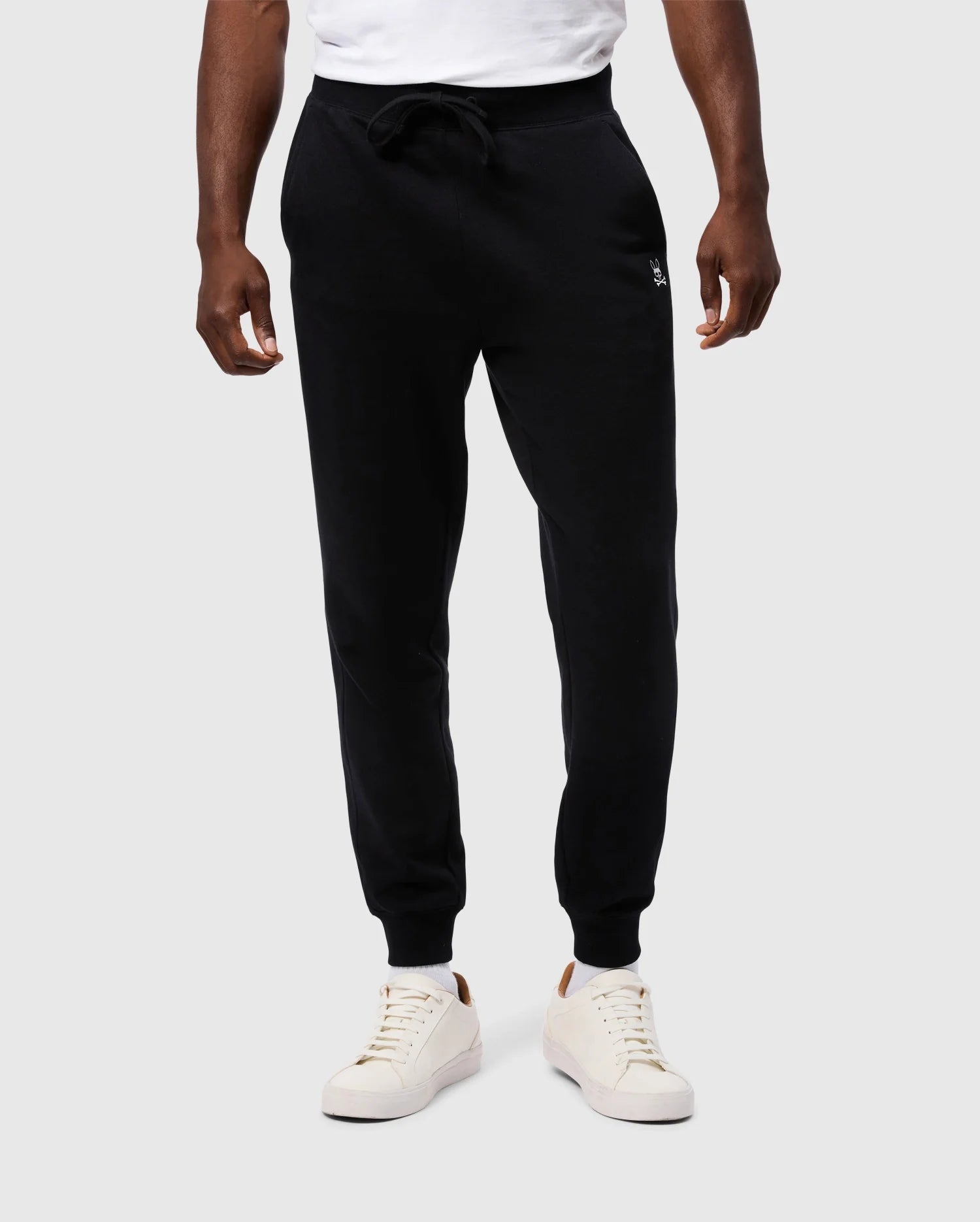 A person is wearing Psycho Bunny's MENS LEVY ESSENTIAL FLEECE SWEATPANT - B6P721D200 in black, paired with a white t-shirt. Known for their comfortable feel, the joggers feature a drawstring waist and fitted ankles. The outfit is completed with white sneakers that have a brown patch on the heel. The background is plain light gray.