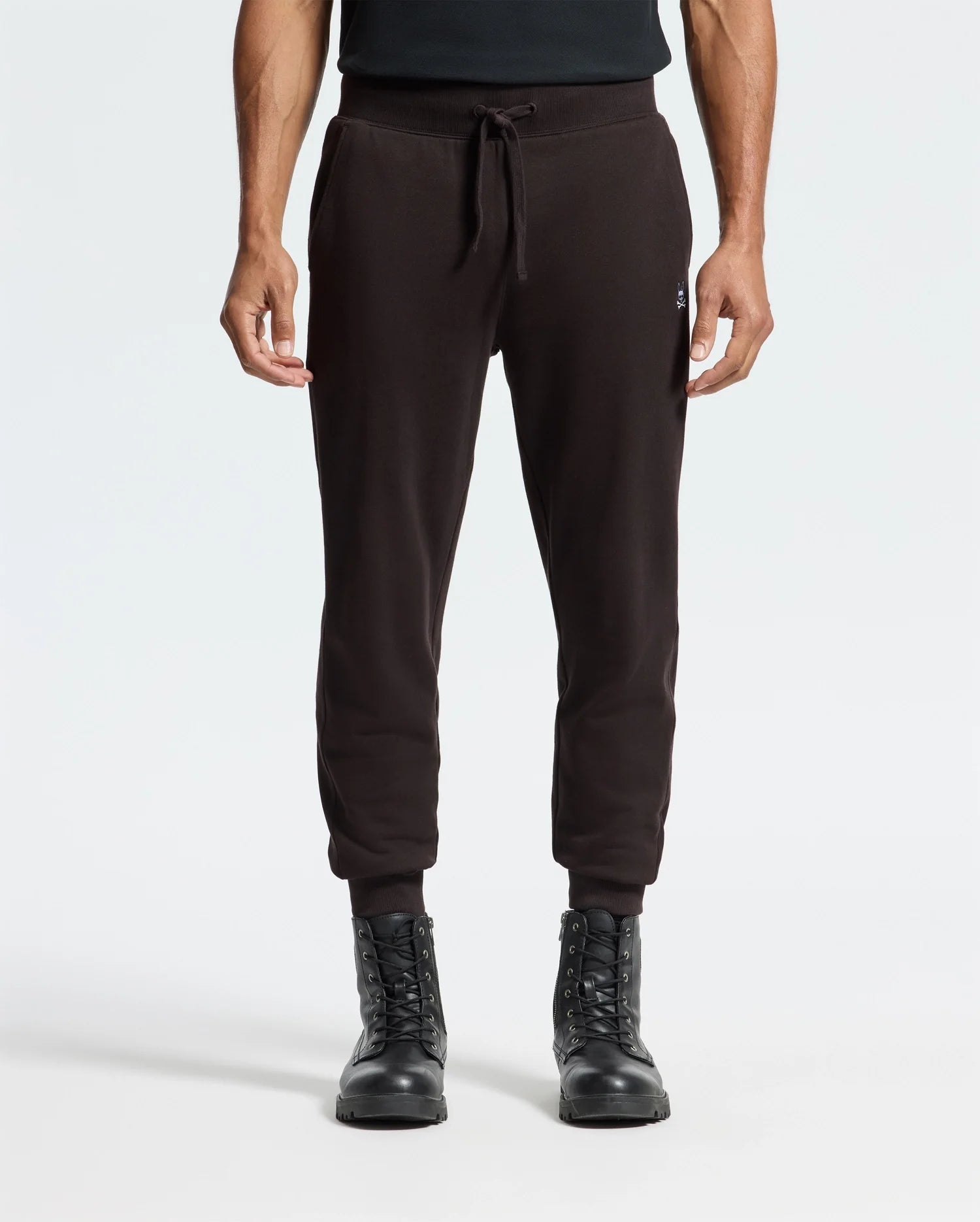 A person wearing Psycho Bunny's MENS LEVY ESSENTIAL FLEECE SWEATPANT (B6P721D200), made from 100% cotton, paired with black lace-up boots, standing against a plain white background.