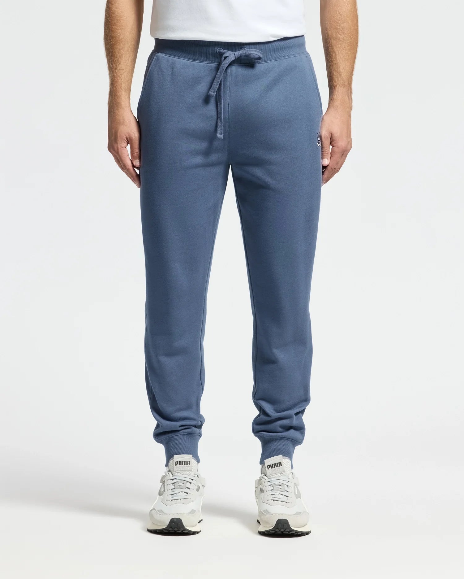 A person wearing the MENS LEVY ESSENTIAL FLEECE SWEATPANT by Psycho Bunny and white Puma sneakers stands against a plain background. The blue joggers, crafted from 100% cotton and featuring a drawstring waist, are ideal for casual wear. The individual is also wearing a white top.