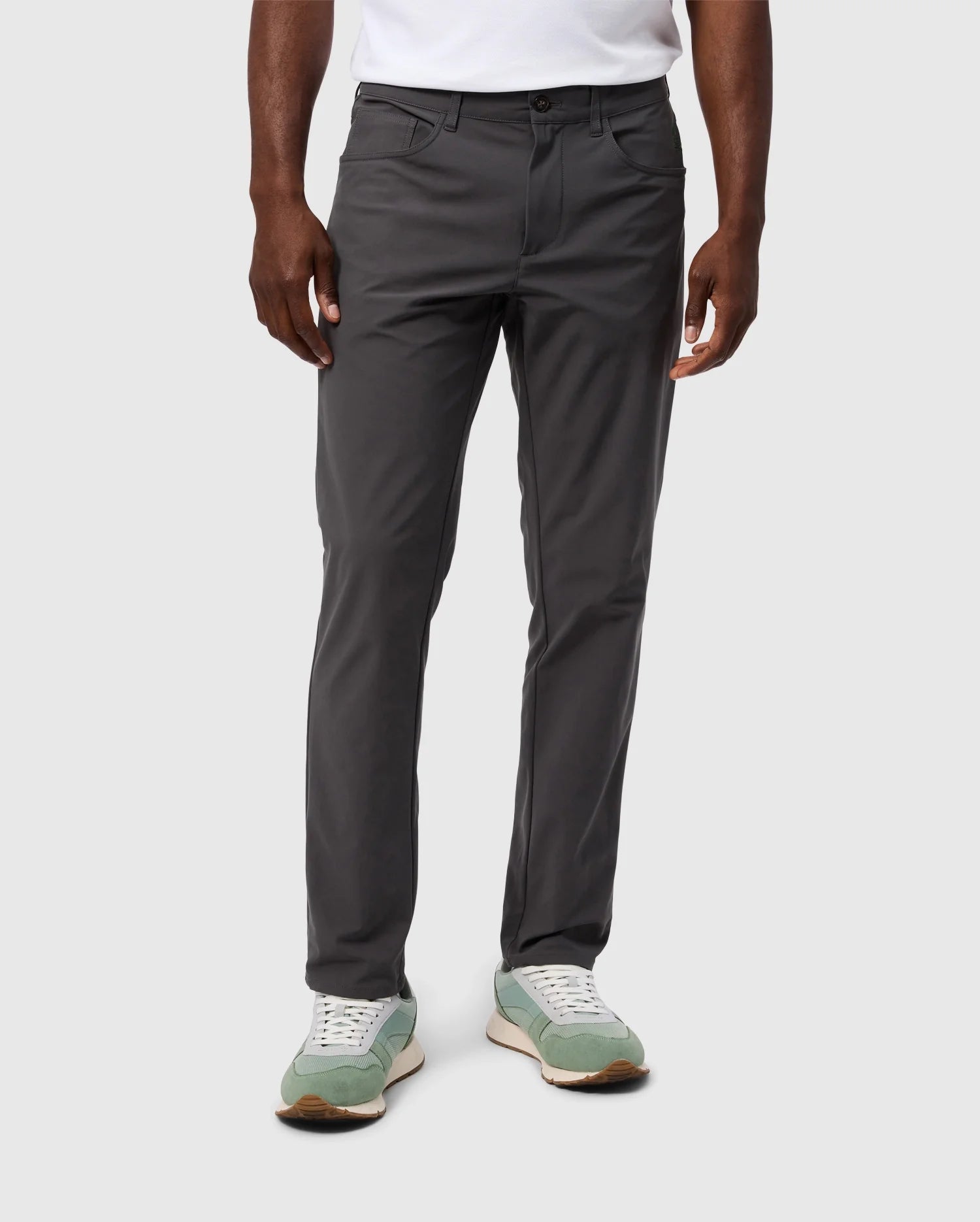 A person is wearing dark gray, slim-fit MENS MAVERICK 10K PANT - B6P788D200 by Psycho Bunny, paired with light green and white sneakers. Their hands are relaxed by their sides, set against a plain light gray background.