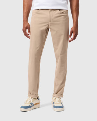 A person is standing wearing Psycho Bunny's MENS MAVERICK 10K PANT - B6P788D200 and white sneakers with blue and beige accents. The background is plain and light-colored, only the lower half of the body is visible.