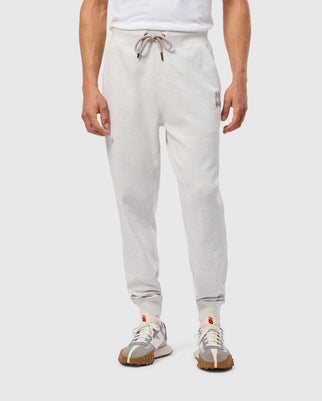 A person dressed in the MENS JAMES PREMIUM SWEATPANT by Psycho Bunny, featuring a light gray cotton-blend fabric with a drawstring waistband and an embroidered design on the upper left leg, stands against a plain backdrop. They wear beige sneakers with brown soles and pair them with a white shirt.