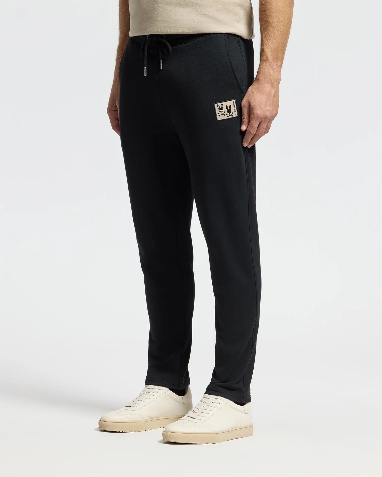 A person is wearing Psycho Bunny's MENS MONROE TWILL PATCH FLEECE SWEATPANT (B6P929D200) in black, featuring a small beige patch on the side, paired with white sneakers. The background is plain white, capturing the view from shoulder height down.