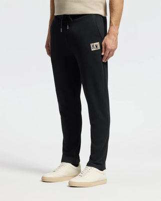 A person is wearing Psycho Bunny's MENS MONROE TWILL PATCH FLEECE SWEATPANT (B6P929D200) in black, featuring a small beige patch on the side, paired with white sneakers. The background is plain white, capturing the view from shoulder height down.