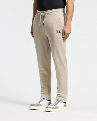 A person wears Psycho Bunny's MENS MONROE TWILL PATCH FLEECE SWEATPANT - B6P929D200 in beige cotton with an embroidered patch, paired with white and beige sneakers. The French terry sweatpants feature a drawstring waist and tapered fit, set against a plain light gray background.