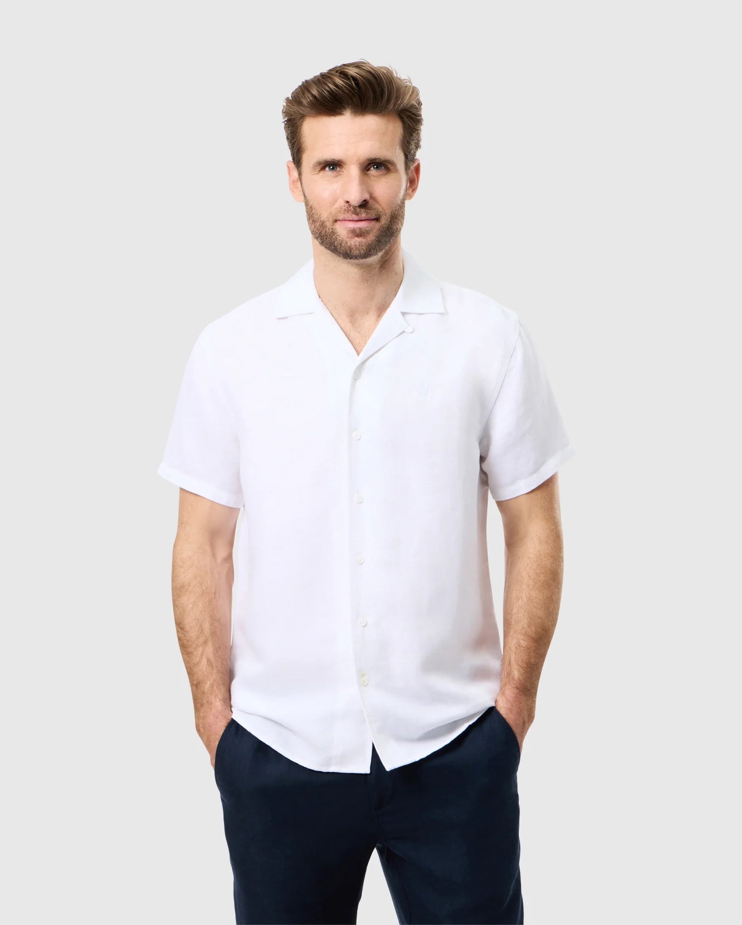 A man with short brown hair and a beard stands against a plain gray background, wearing Psycho Bunny's MENS WINDCREST LINEN SHORT SLEEVE SHIRT - B6Q148C200 in white and dark pants, with his hands in his pockets.