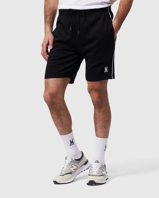 A person is wearing the black, comfort-driven Psycho Bunny MENS LEON REGULAR FIT SWEATSHORT - B6R205B200 featuring a white logo on the left leg, paired with white crew socks adorned with black logos, and gray New Balance sneakers. The background is plain white.