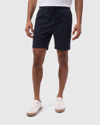 A person wearing dark blue, eco-friendly Psycho Bunny MENS WILLIS STRETCH TENCEL SHORT - B6R239Y1WB with a drawstring waist and a white t-shirt is standing against a plain white background. They are also wearing white sneakers with light brown soles. The image focuses on the person's lower half, from waist to shoes.