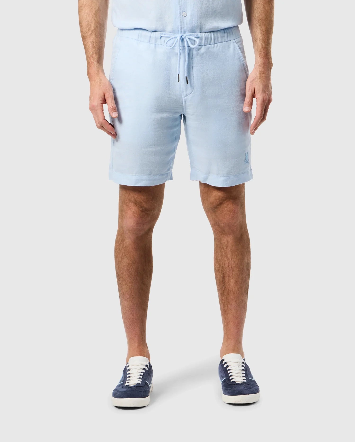 A man wearing MENS WINDCREST LINEN SHORT - B6R474C200 by Psycho Bunny and dark blue sneakers stands against a plain background, showing only his lower torso and legs.