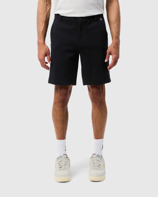 A person stands facing forward, wearing a pair of Psycho Bunny MENS SHIRO KNIT HONEYCOMB SHORT - B6R567C200 made from sweat-wicking knit honeycomb fabric. Complementing the shorts are white socks with a black logo and light-colored sneakers. The background is plain white, and the model's hands rest by their sides.