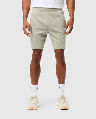 A person is standing against a plain background, wearing light green MENS LYONS SWEATSHORT - B6R578C200 by Psycho Bunny with pockets, a white shirt, white socks, and beige sneakers. The stance is relaxed with arms hanging down by their sides. Only the lower half of the body is visible in the image, showcasing the embroidered Bunny appliqué on the shorts.