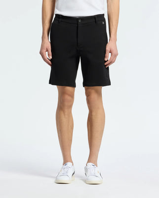 A person wears black Psycho Bunny MENS SHIRO KNIT HONEYCOMB SHORT - B6R585F200, a white shirt, and white sneakers. The plain white background highlights the honeycomb knit texture of the shorts, with their arms by their sides.