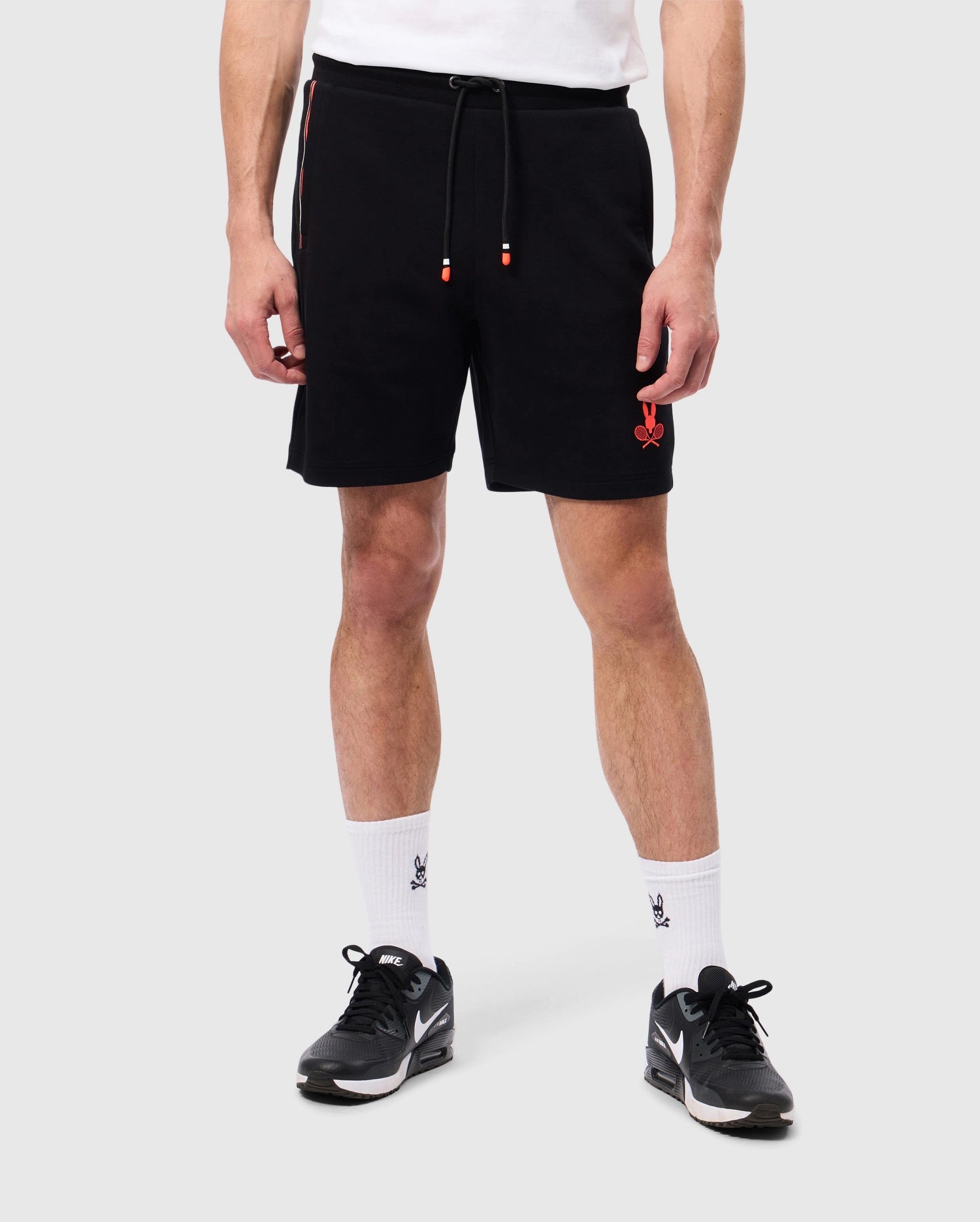 A person is wearing Psycho Bunny's black MENS COURTSIDE TRACK SHORT - B6R690C200, which features a red emblem near the left hem, made from performance-ready fabric. They have paired the shorts with white socks that feature a black emblem and black sneakers. Their hands are relaxed by their sides, set against a plain gray background.