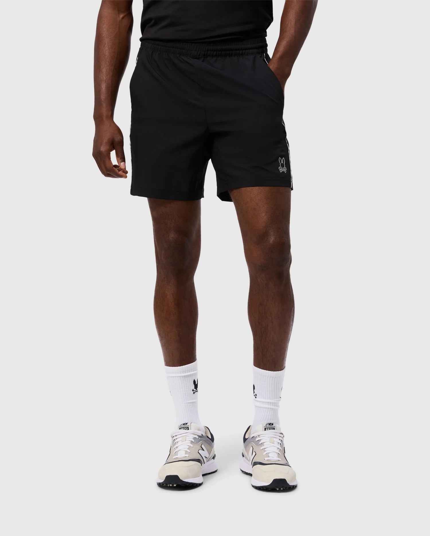 A person wearing a black MENS MANSFIELD SPORT SHORT by Psycho Bunny, paired with a black T-shirt, white socks, and light-colored sneakers stands against a plain background. The individual's face is not visible, and their stance is casual.