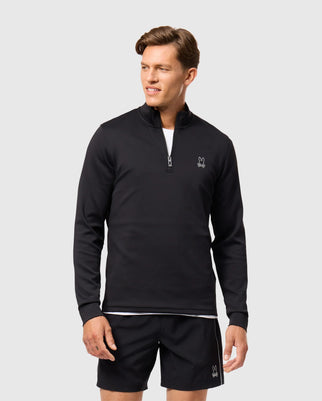 A man in Psycho Bunny's MENS LAURIER 1/4 ZIP HOODIE B6S149D200 and matching black shorts stands against a plain backdrop. With an athleisure vibe, he gazes left with a neutral face. The outfit features subtle embroidered logos on both the hoodie and shorts.